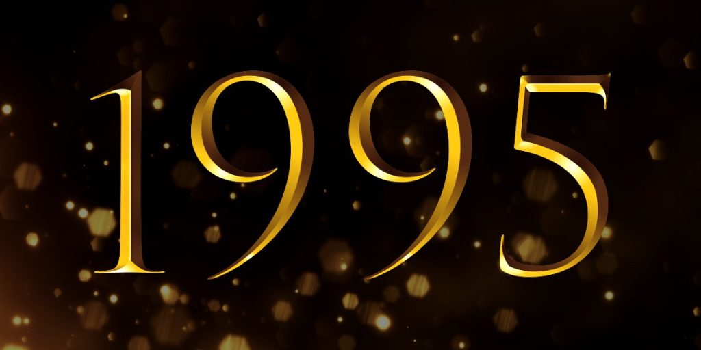 A group of elegantly dressed individuals stands together, smiling, with a red carpet and awards in the background.