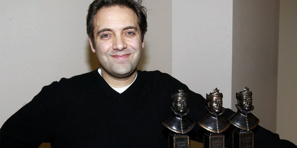 A man in a black sweater smiles while holding two Olivier Awards statues.