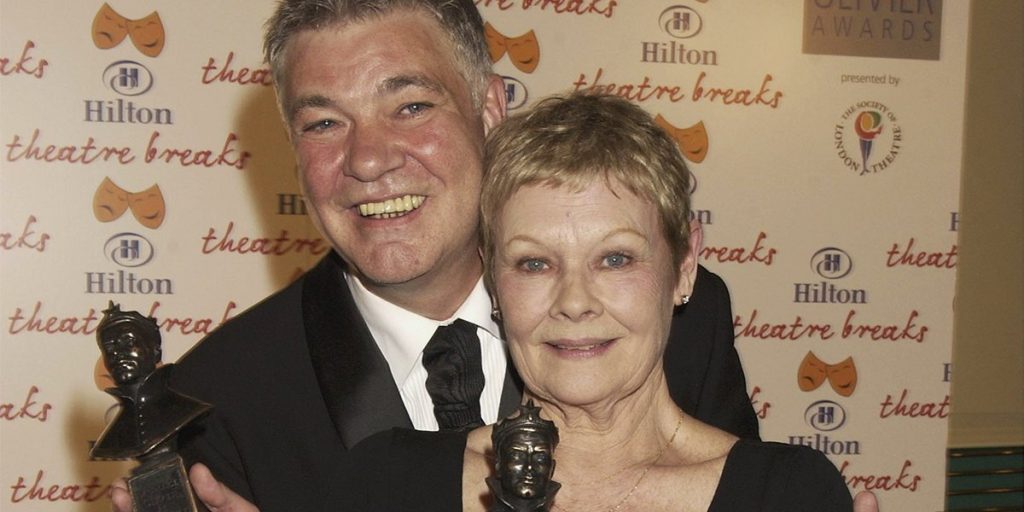 Judi Dench and a man smile together, both dressed formally, against a backdrop of event signage.