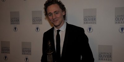 A man with curly hair in a black suit holds an award, smiling against a white backdrop.