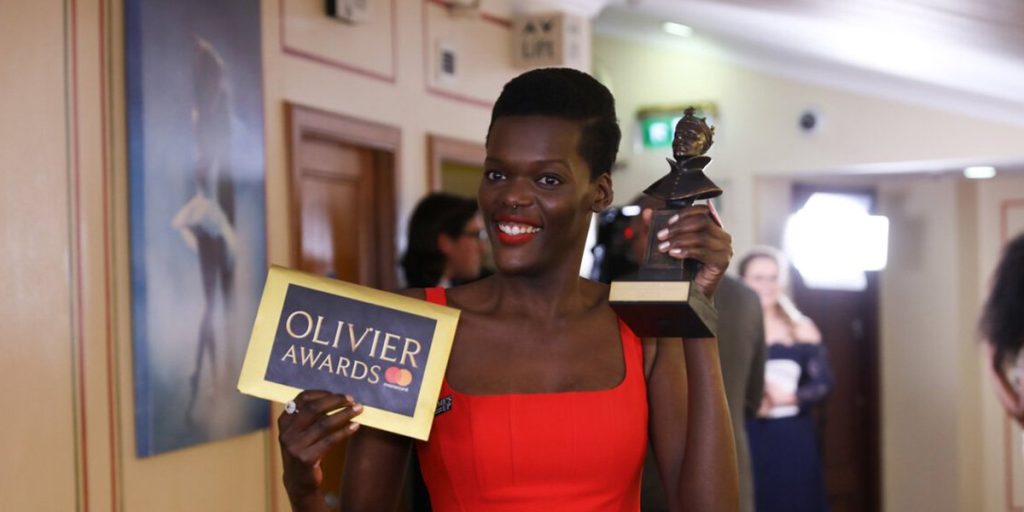 A woman in a red dress smiles, holding an Olivier Award statue and a sign reading "Olivier Awards.