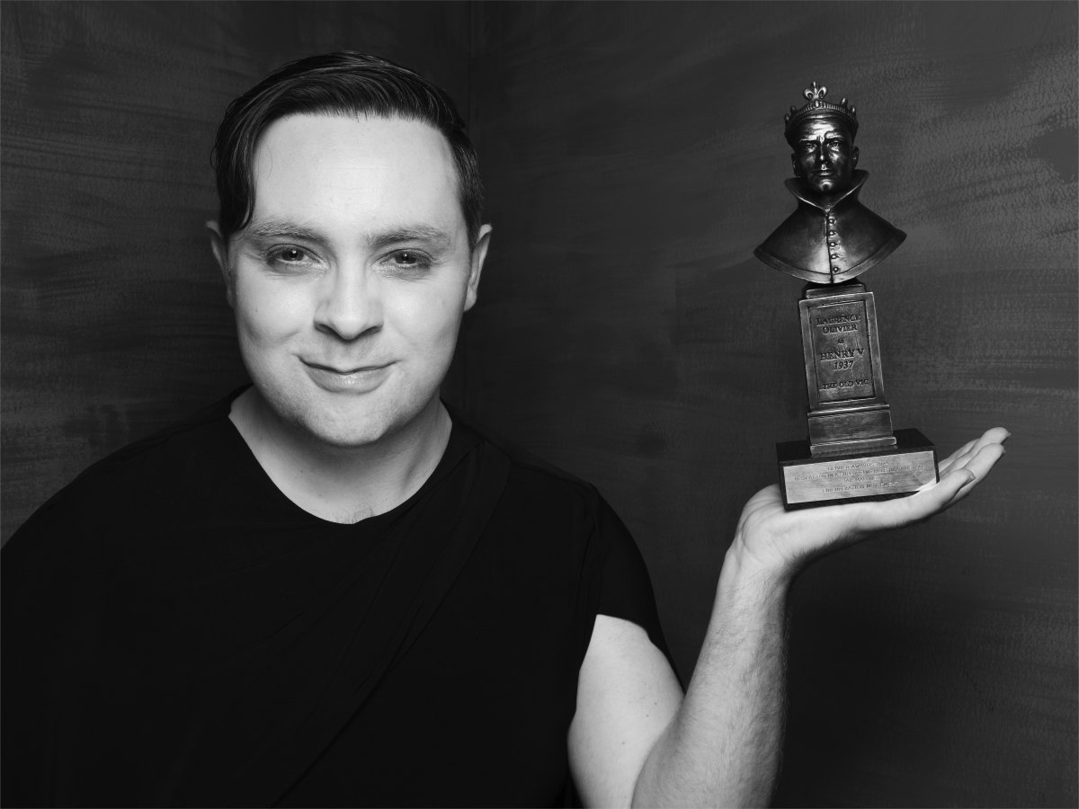 A person smiling while balancing an Olivier Award statue on the palm of their hand.