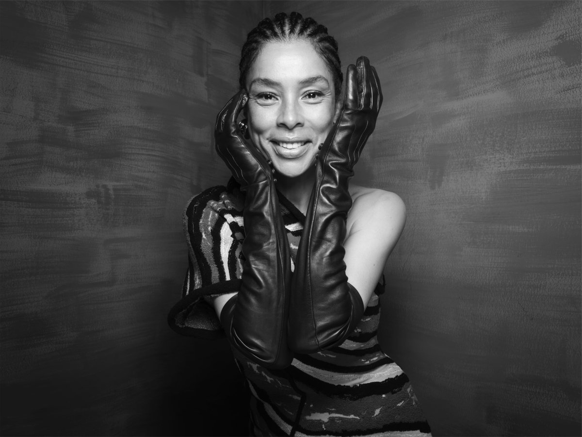 A woman wearing a dress and long leather gloves. She is smiling and her hands are gently cupping her face.