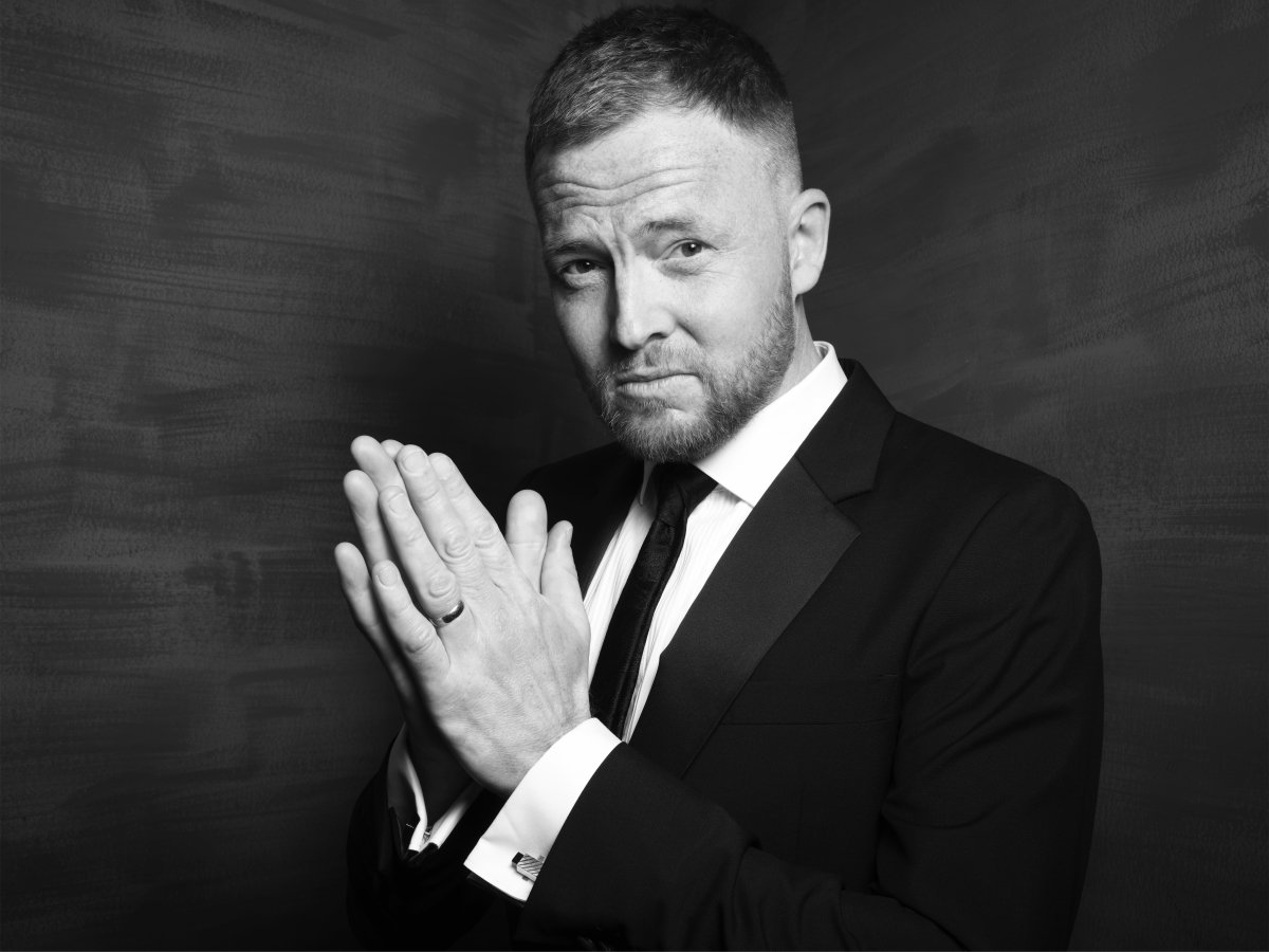 A man in a suit. He has his hands together in a praying motion. He is standing slightly angled away from the camera.