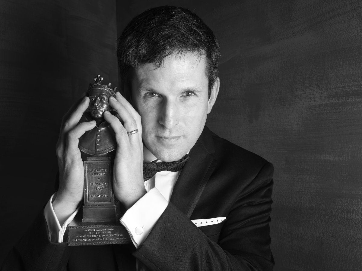 A man in a shit and bow tie. He is holding an Olivier Award in both hands. He is holding the statue up next to his face and looking intently at the camera.