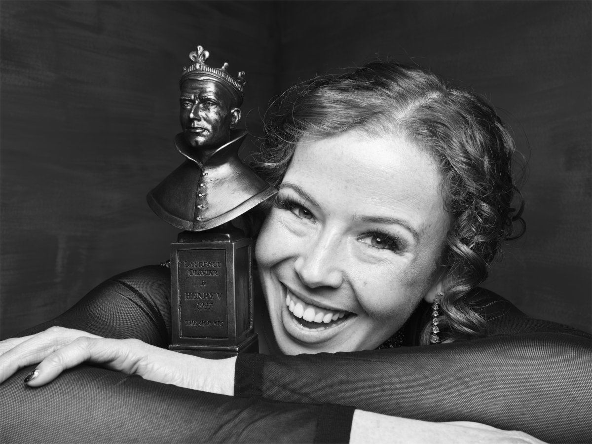 A woman smiling with her arms crossed. She is leaning her face on her arm and hugging an Olivier Award statue.