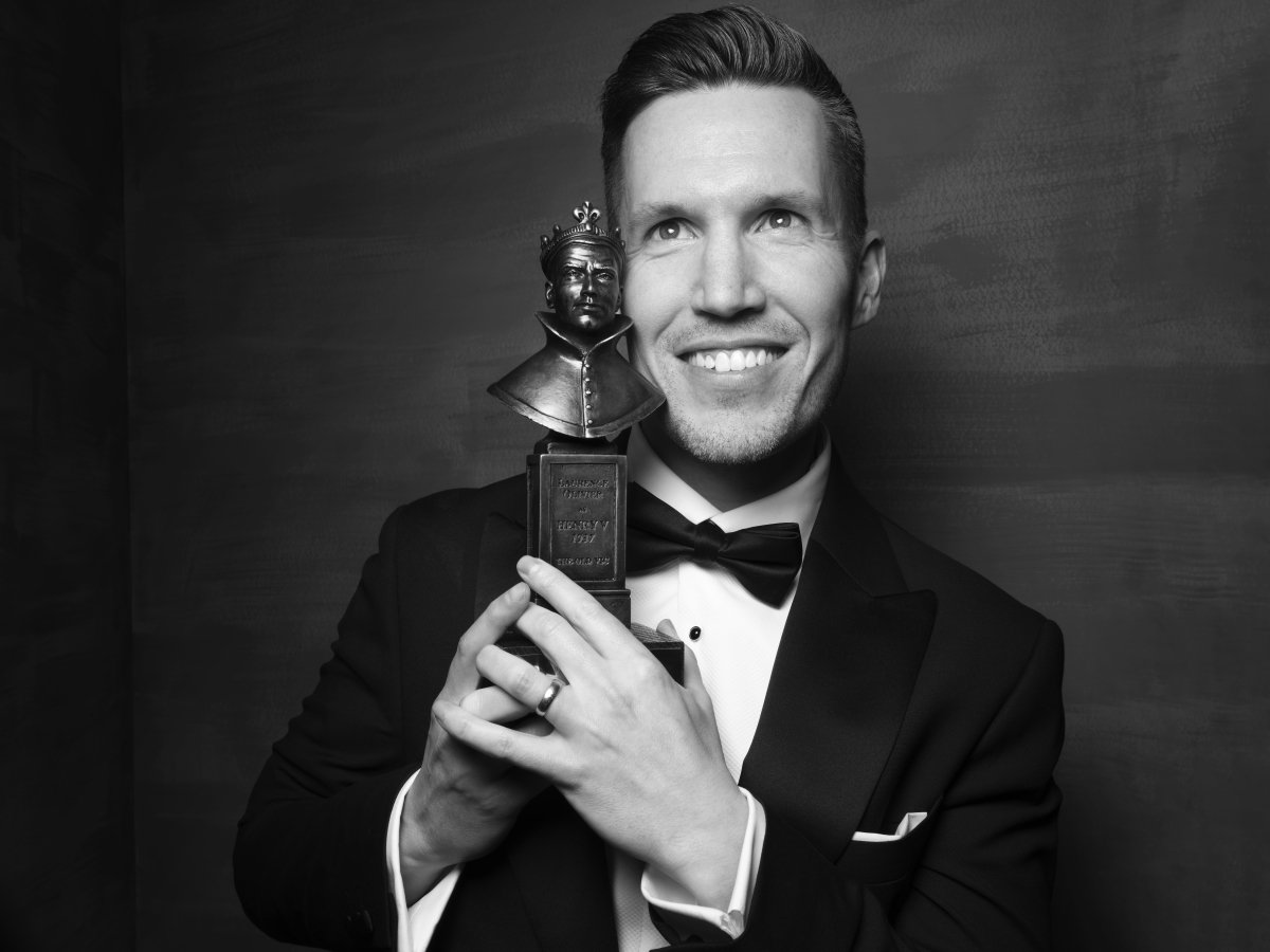 A man wearing a suit and bow tie. He is holding an Olivier Award statue up next to his face and looking up and smiling.