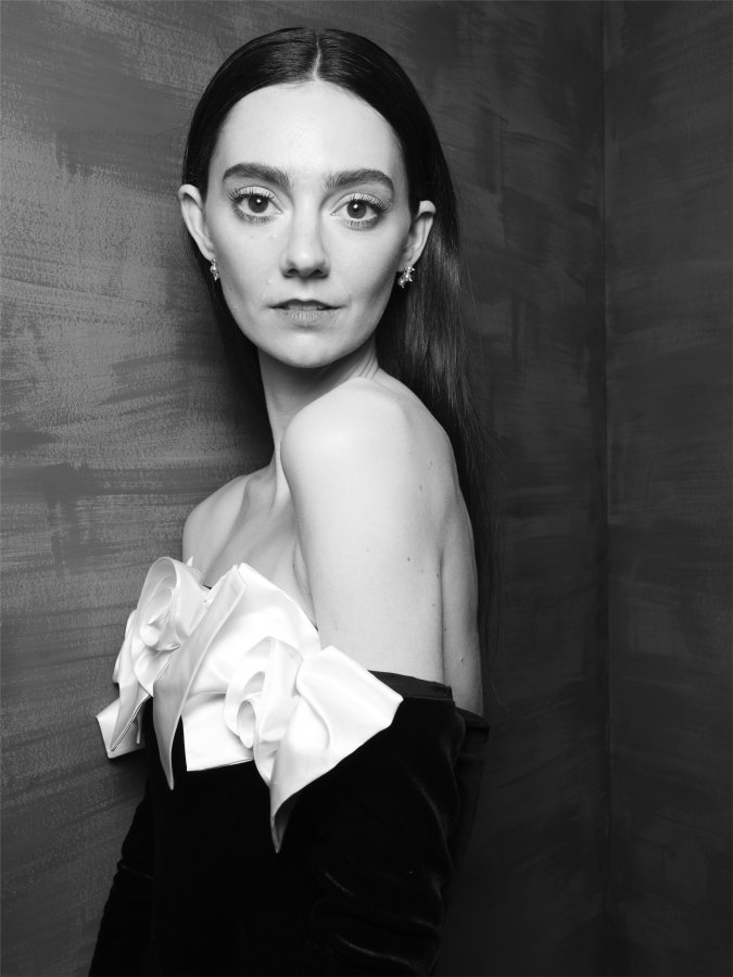 A woman standing side on but with her head facing the camera. She is wearing a black dress with white frills on the neckline.