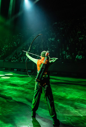 A performer in an orange top draws a bow, surrounded by green lighting and an audience in the background.