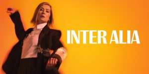 The background is yellow which fades into orange towards the edges. On the left is a picture of a person with shoulder length blonde hair, they are wearing a black blazer and white shirt. They have their right arm up in the air and their left arm out front. On the left it reads 'Inter Alia'