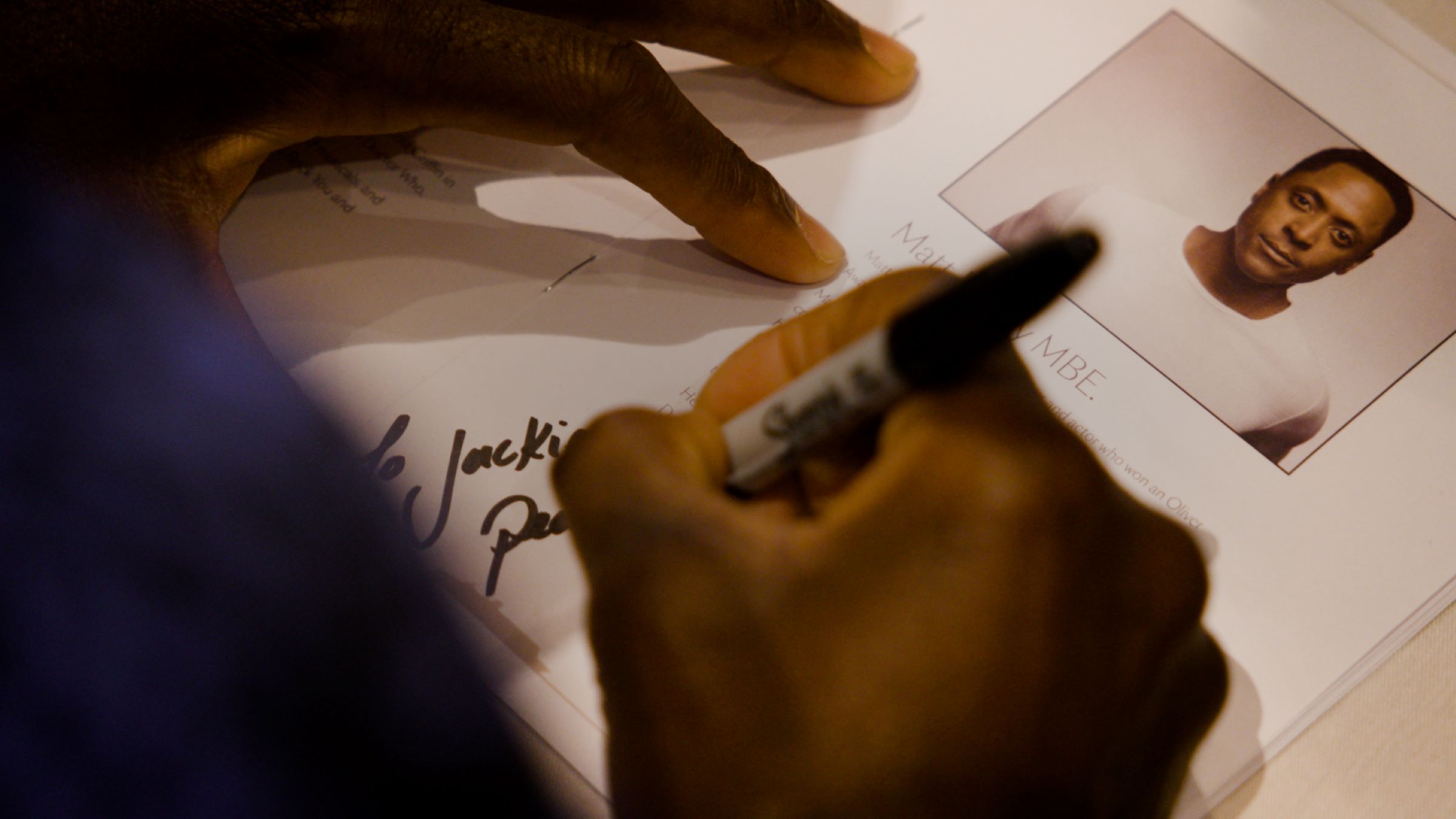 A hand writes on a paper with a black marker, signing a name.