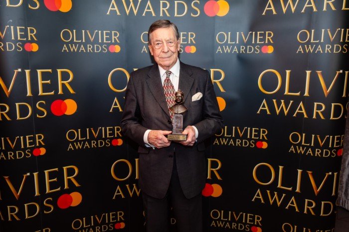 Richard Walton holds his Olivier Award statuette in front of a branded backdrop