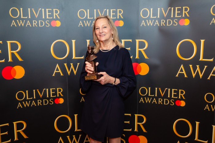 Susan Whiddington holds her Olivier Award statuette in front of a branded backdrop