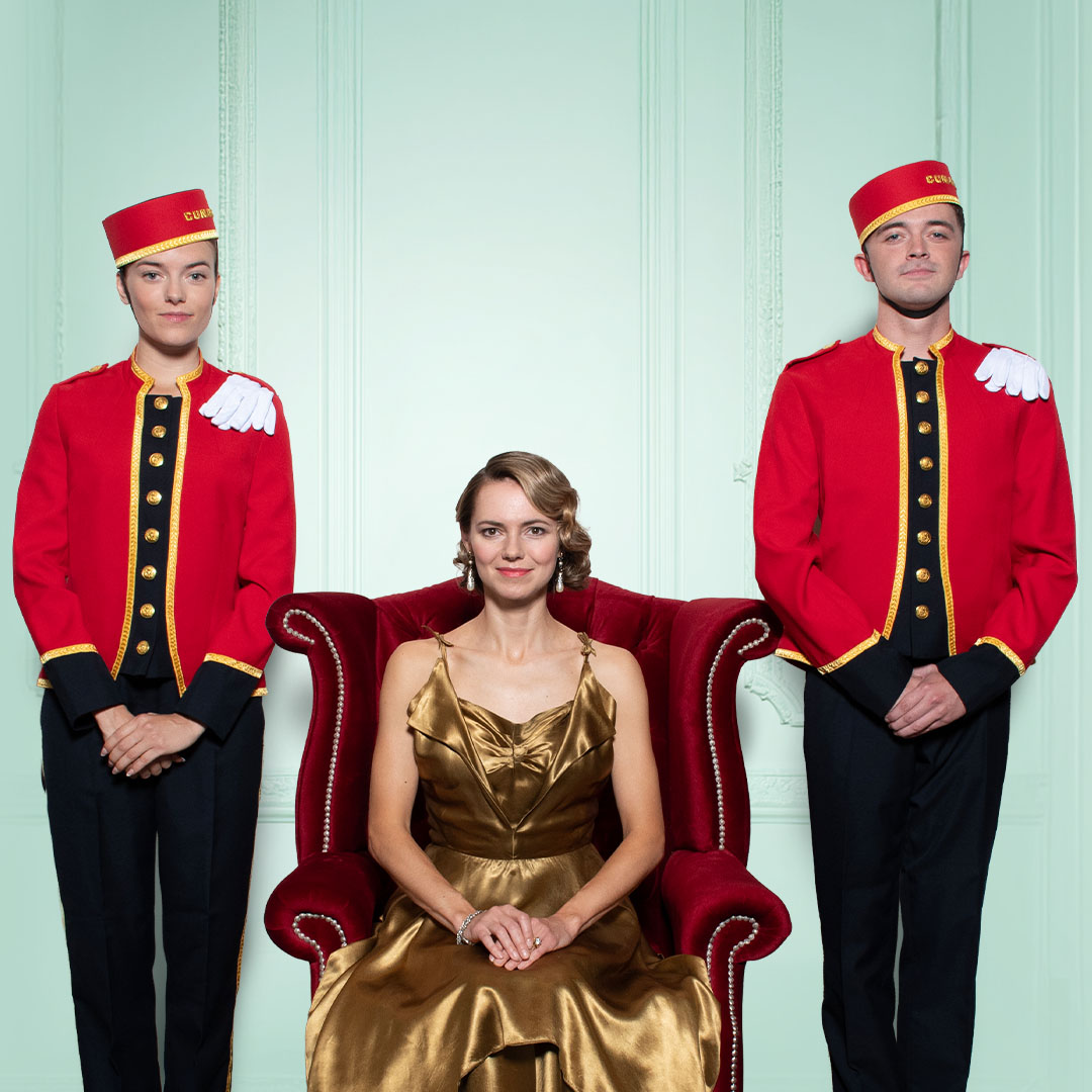 A group of elegantly dressed bellhops stands in a row, smiling against a decorative backdrop for the Olivier Awards.