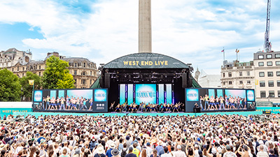 An image of an audience watching the cast of Mamma Mia perform at West End LIVE. The stage is set in Trafalgar Square with blue sky and a statue behind the stage.