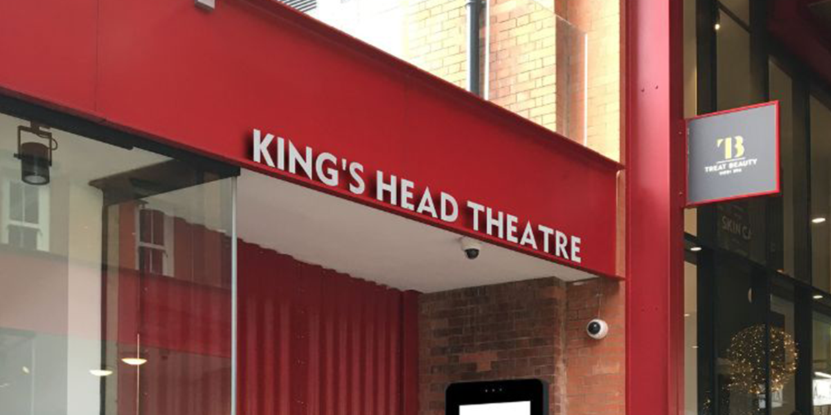 A performer in a vibrant costume gestures expressively on stage, with dramatic lighting and a dark background.